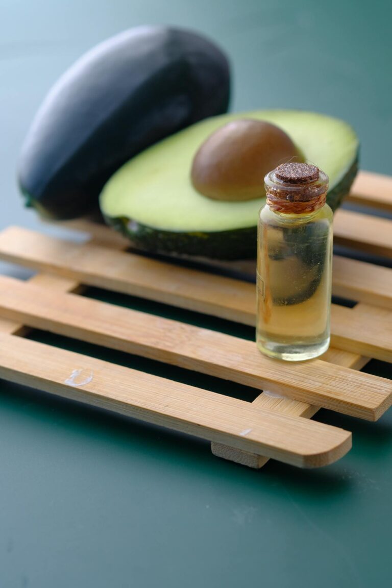 Glass bottle of avocado oil serum beside a sliced avocado on a bamboo mat.